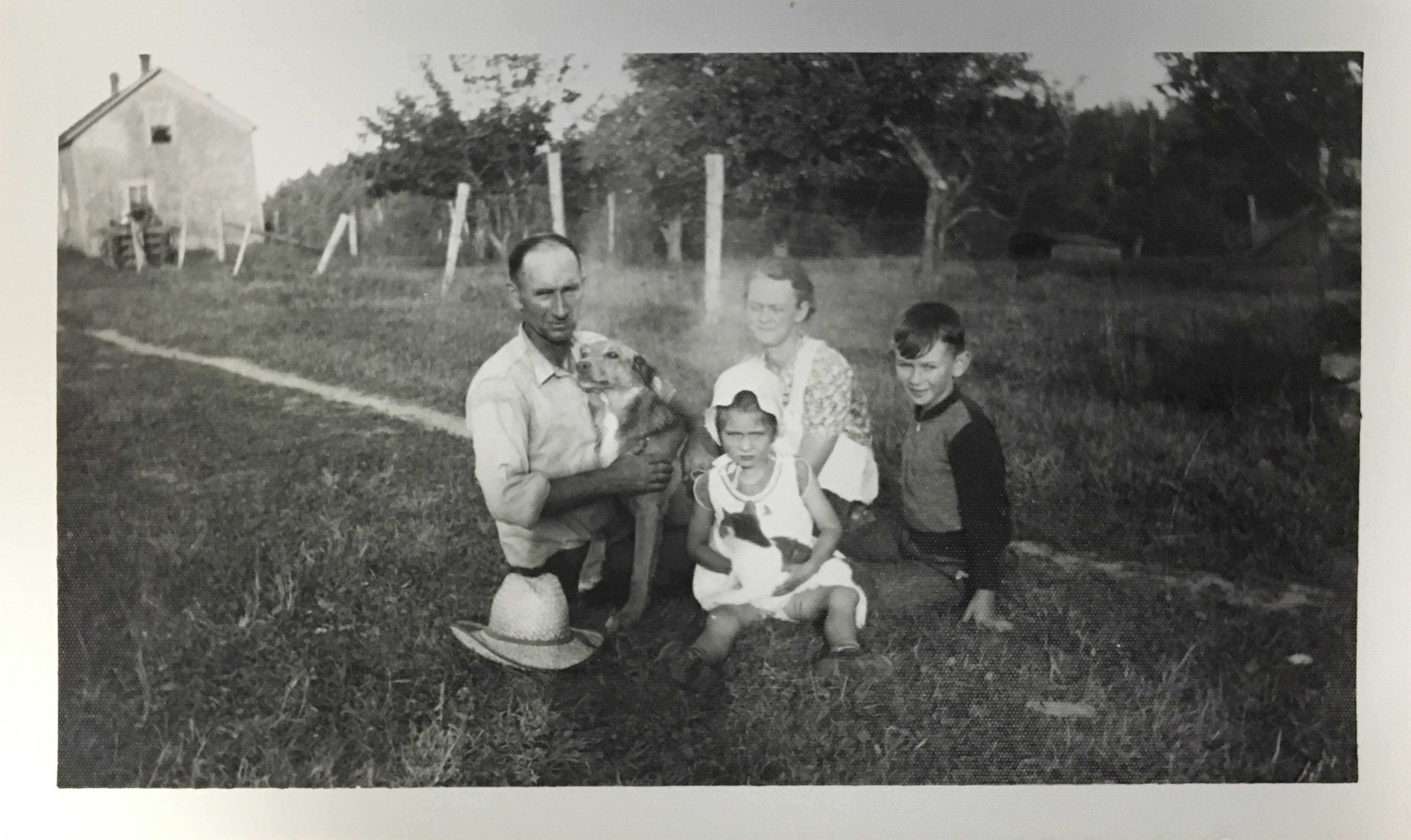 Black and white photograph. Archie and his family sit on the lawn of their farm. Archie holds on to the family dog Brownie. Daughter Margie, wife Grace, and son Francis are to his left.
